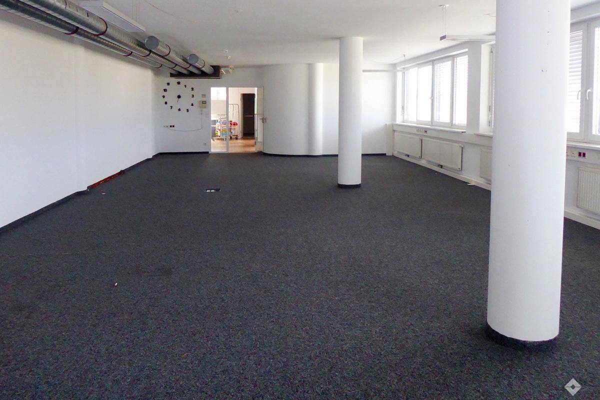 Weitläufiger Bürobereich mit dunklem Teppichboden, Säulen und großen Fenstern, die viel Tageslicht hereinlassen.