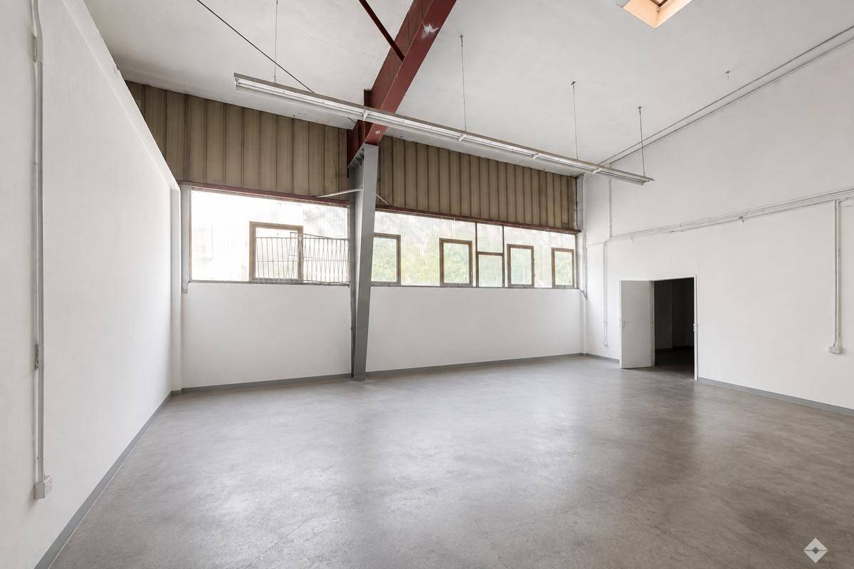 Große, leere Industriehalle mit hohen Decken, Betonboden und vielen Fenstern, die Tageslicht hereinlassen.