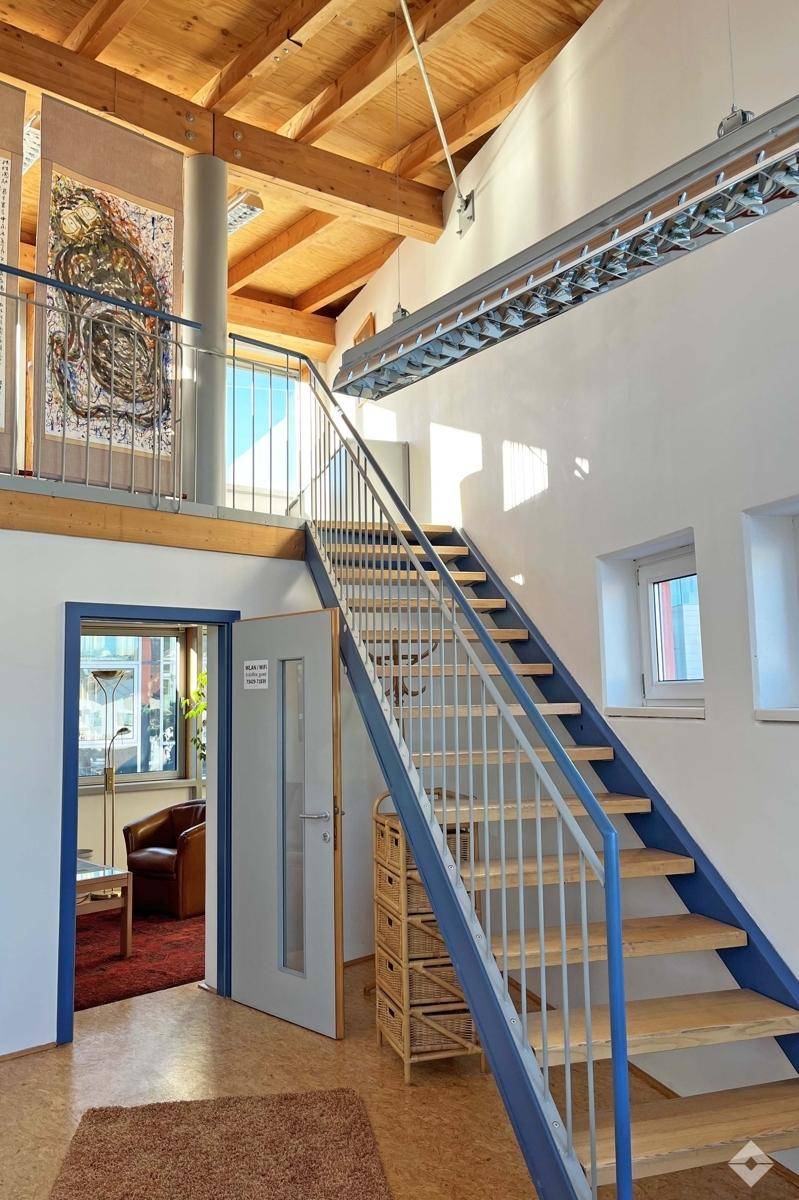 Hoher Eingangsbereich mit offener Holztreppe, Holzbalkendecke und Galerie mit Kunstwerken.