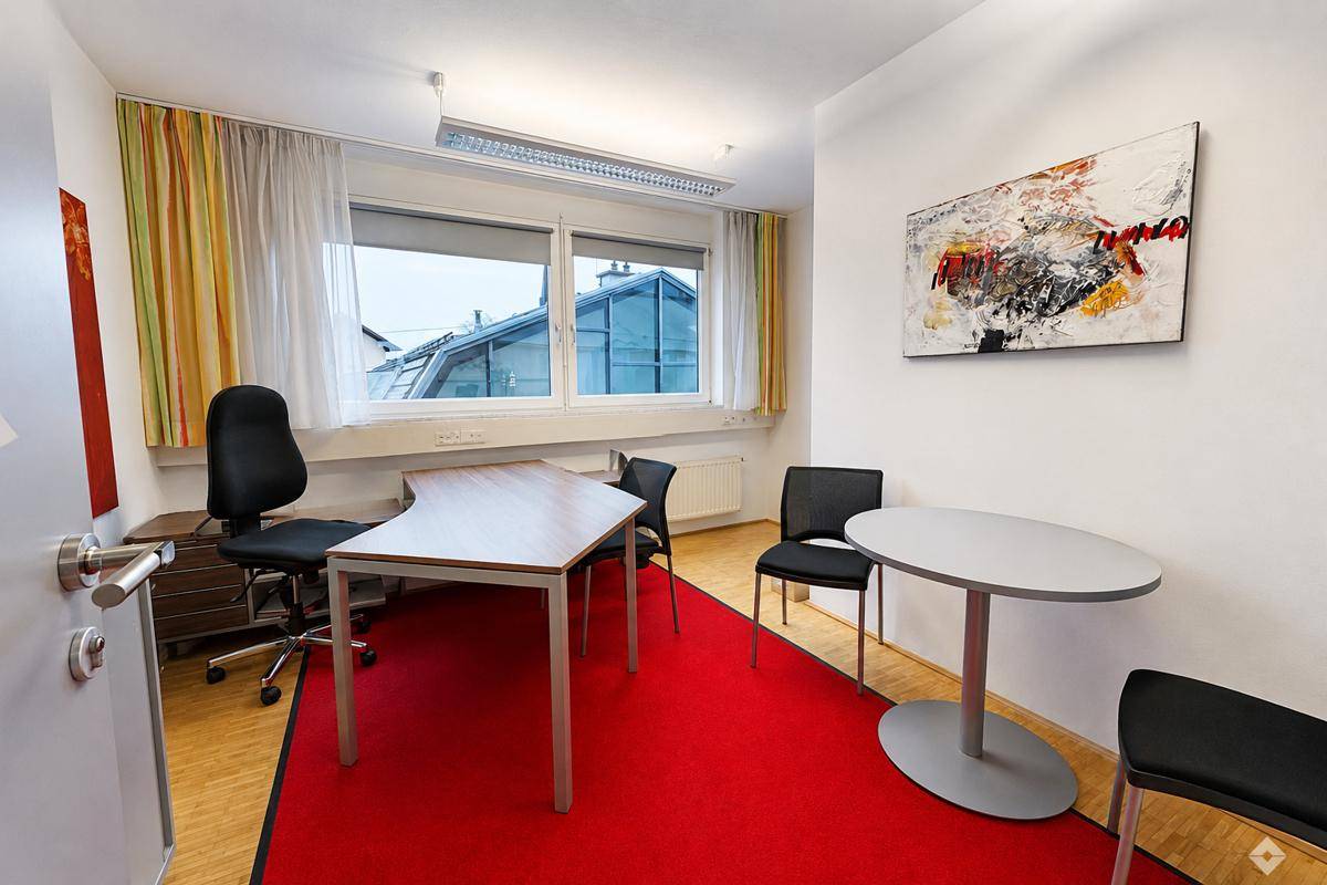 Ein helles Büro mit rotem Teppich, Holzboden und einem Schreibtischbereich mit Blick nach draußen.