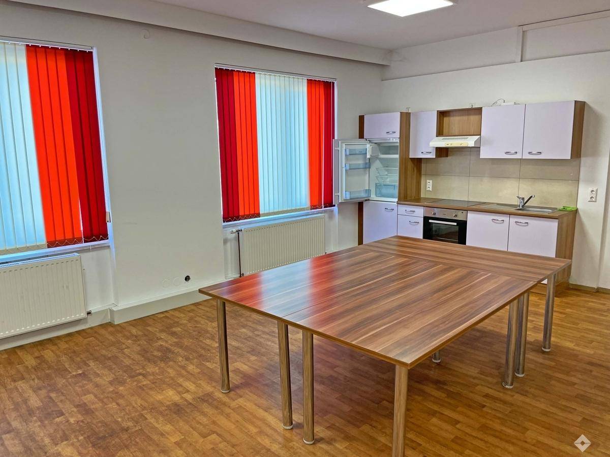 Funktionale Büroküche mit kleiner Küchenzeile, großem Holztisch und Fenstern mit roten Jalousien.