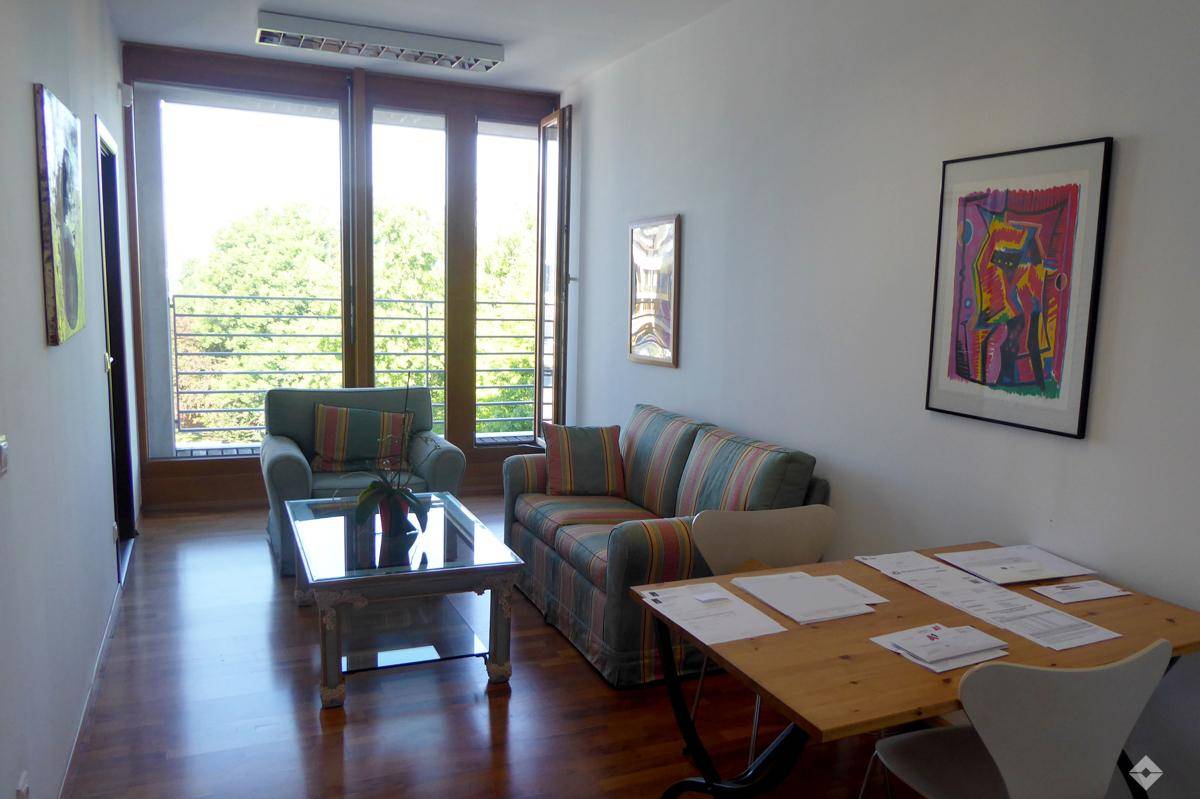 Gemütlicher Wohnbereich mit Sofa, Sessel und einem Schreibtisch, umgeben von großen Fenstern mit Grünblick.