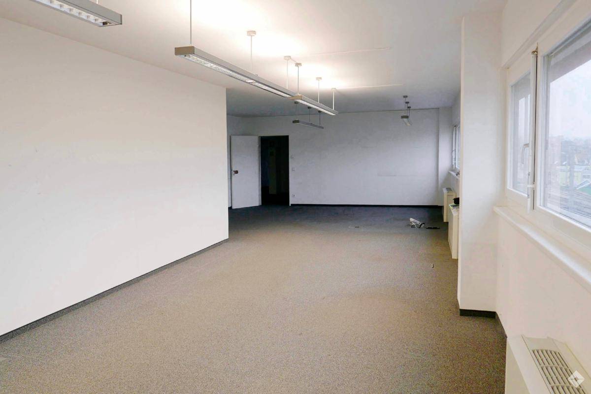Leerer Büroraum mit hellem Teppichboden, weißen Wänden und einer Tür, die zu einem weiteren Bereich führt.
