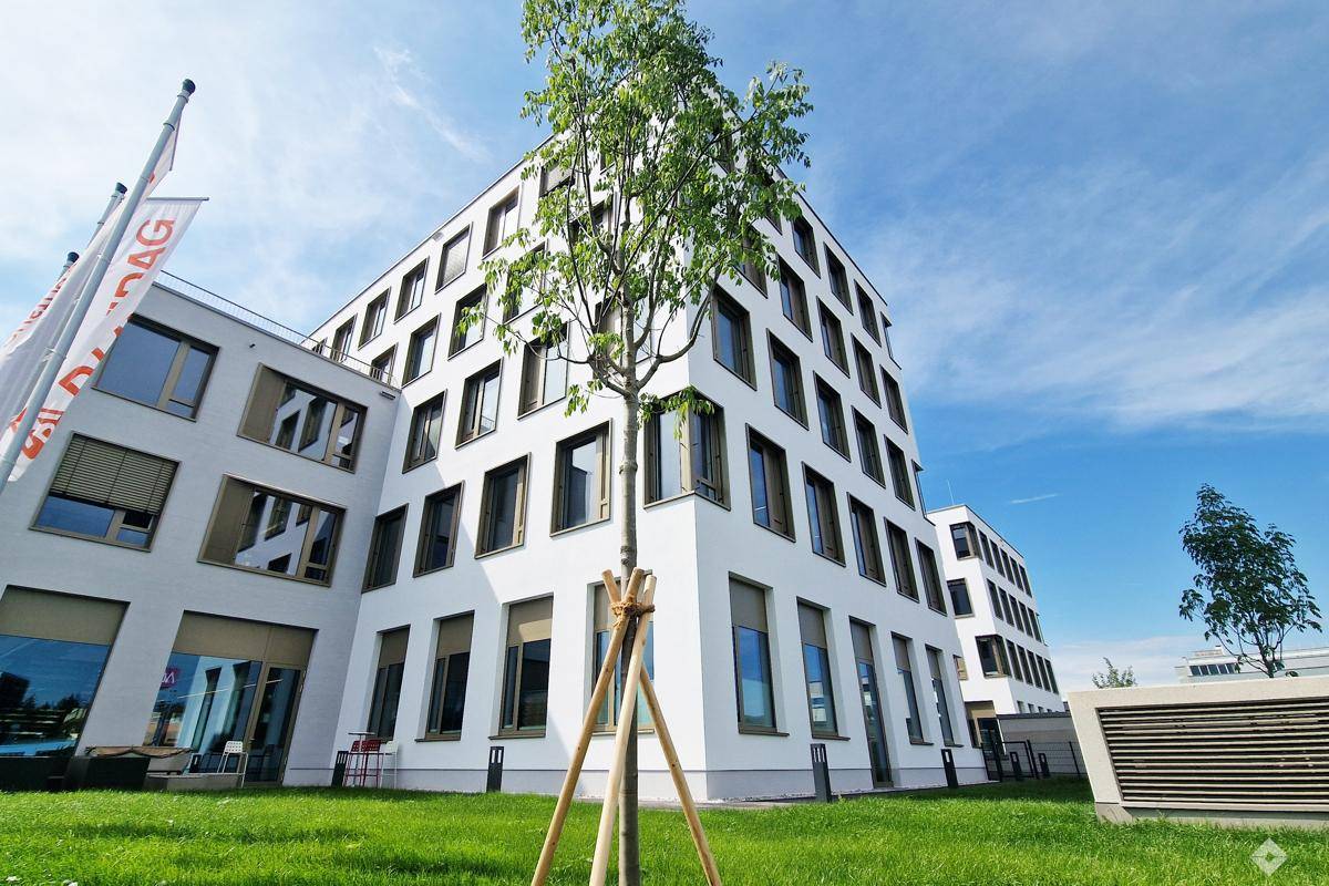 Modernes Bürogebäude mit weißer Fassade, großen Fenstern und gepflegter Grünfläche unter blauem Himmel.