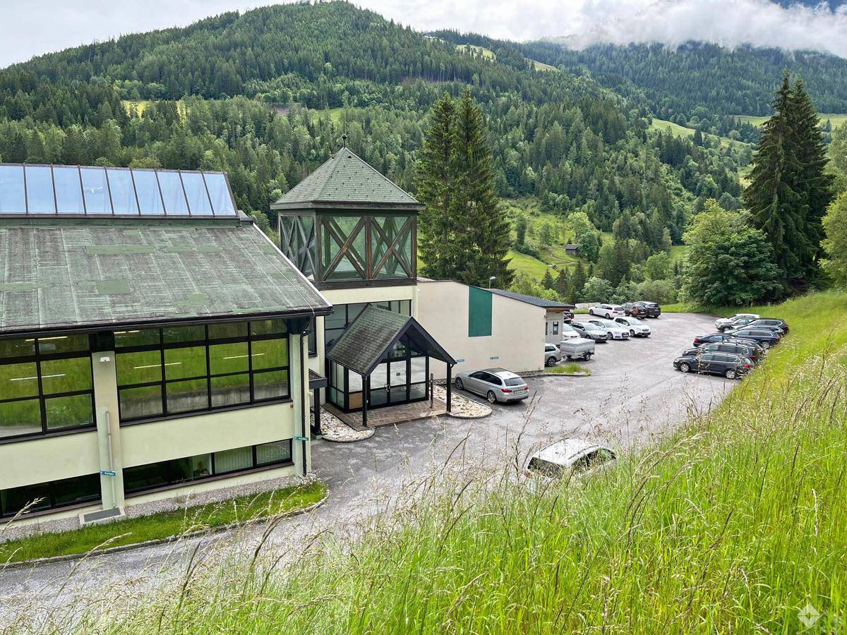 Seitenansicht des Gebäudes mit großem Parkplatz und der beeindruckenden Berglandschaft im Hintergrund.
