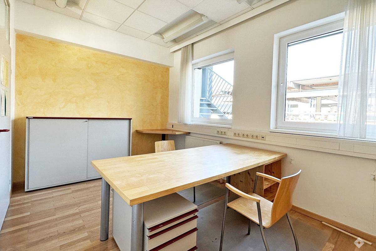 Kleineres Büro mit Holztisch, gelber Akzentwand und zwei Fenstern für ausreichend Tageslicht.