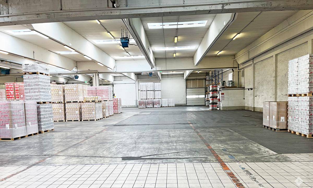 Große Lagerfläche mit unterschiedlichen Bodenbelägen, hohen Decken und gestapelten Waren, ideal für Logistik.