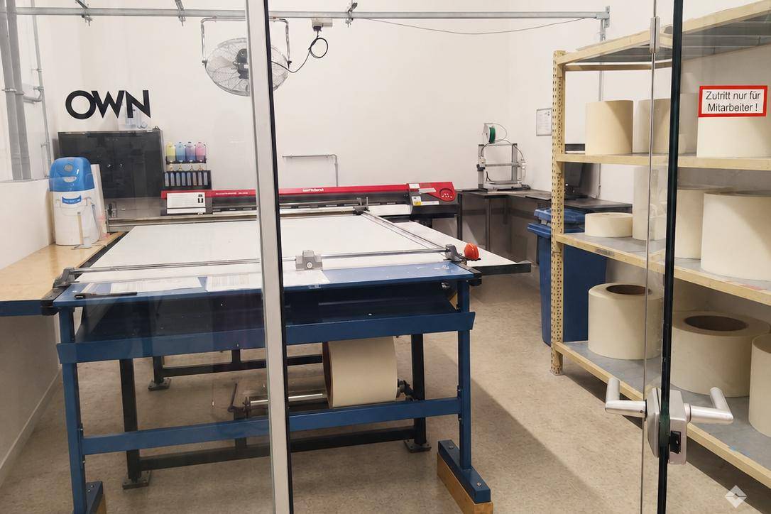 Produktionsraum mit Großformatdrucker, Schneideplotter und Materialrollen, sichtbar durch eine Glastür.