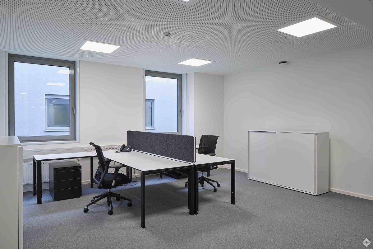 Helles Büro mit zwei Arbeitsplätzen, modernen Schreibtischen und einem Aktenschrank, gut beleuchtet durch Fenster.