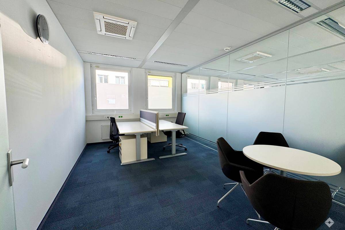 Modern eingerichtetes Büro mit blauer Teppichboden, Glaswänden und mehreren Arbeitsbereichen für effizientes Arbeiten.