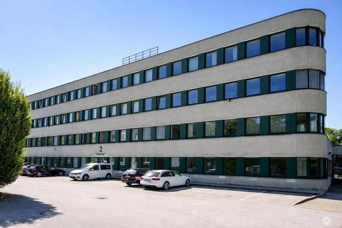 Modernes Bürogebäude mit abgerundeten Ecken und zahlreichen Fenstern, umgeben von Parkplätzen unter blauem Himmel.