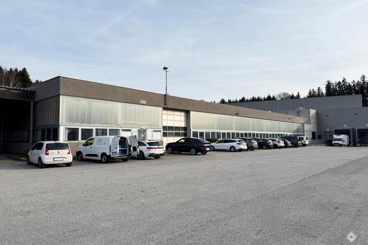 Weitläufige Außenansicht eines Industriegebäudes mit großem Parkplatz und mehreren Fahrzeugen unter blauem Himmel.