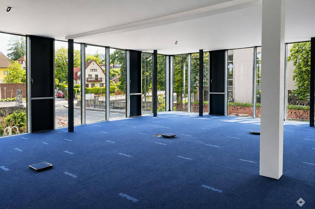 Geräumiger, leerer Büroraum mit blauem Teppichboden und großen Fenstern mit Blick ins Grüne.