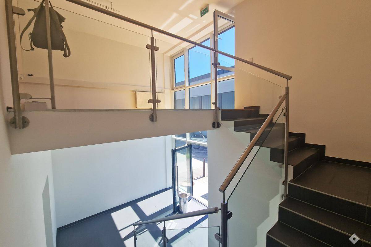 Modernes Treppenhaus mit dunklen Stufen, Glasgeländer und viel Tageslicht durch große Fenster.