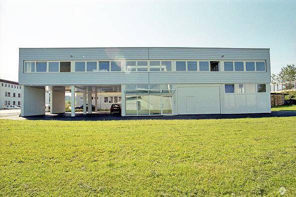 Modernes Geschäftsgebäude mit großer Rasenfläche und überdachtem Parkbereich im Erdgeschoss.