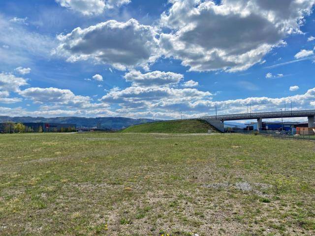 Großes offenes Feld mit einer Brücke im Hintergrund, unter einem weiten Himmel mit Wolken und Bergsicht.