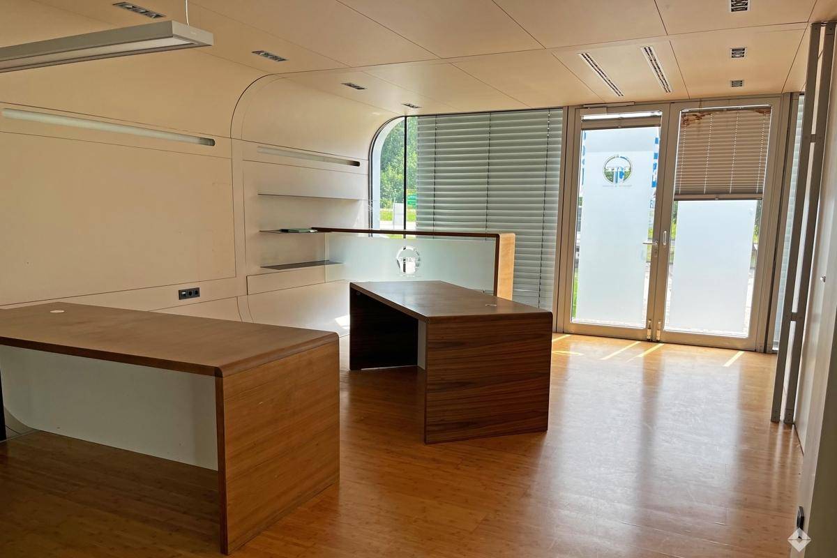 Moderner Büroraum mit Holzboden, zwei Schreibtischen und großen Fenstern mit Jalousien.