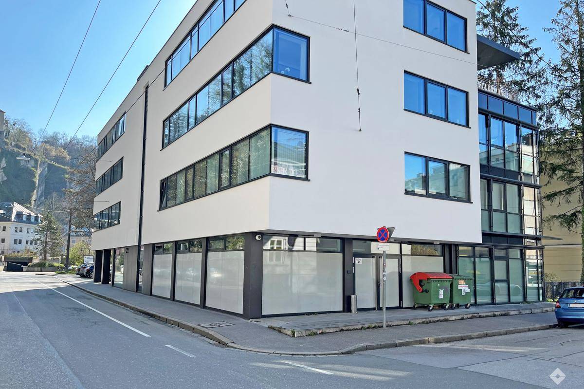 Modernes Bürogebäude an einer Straßenecke mit weißer Fassade und großen Fenstern, umgeben von Bäumen.