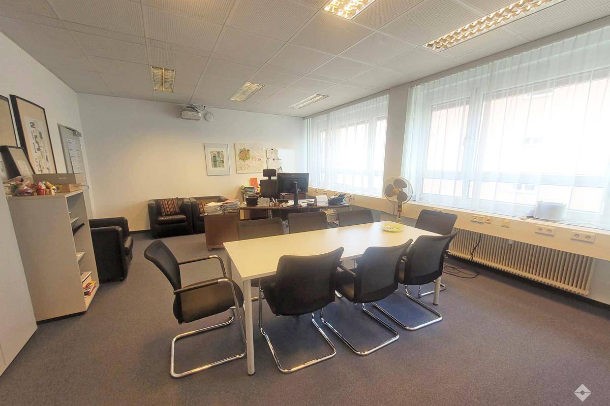 Geräumiges Büro mit Konferenztisch, Teppichboden und großen Fenstern, die viel Tageslicht hereinlassen.