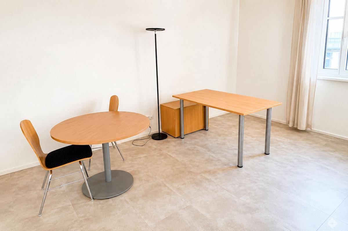 Kleiner Bürobereich mit Schreibtisch, ovalem Tisch und Stühlen auf hellem Fliesenboden.
