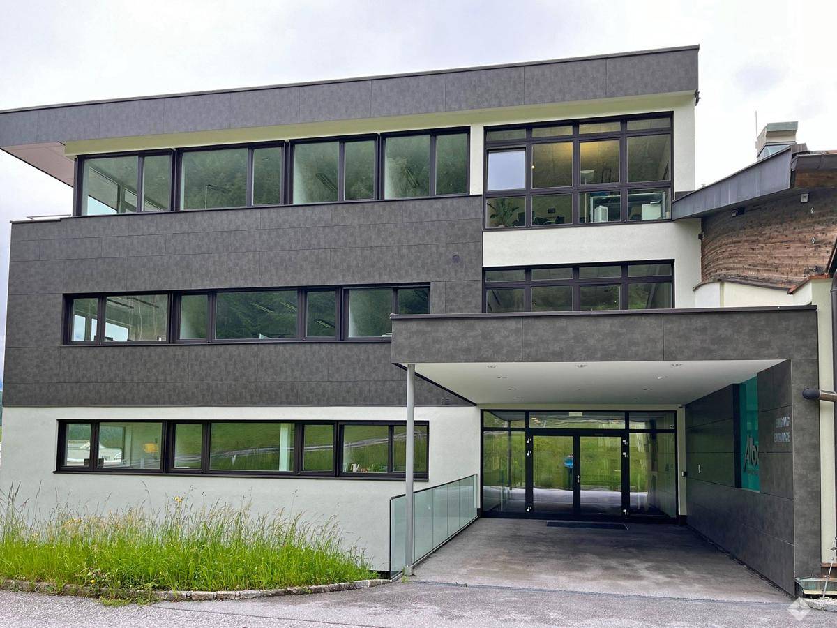 Moderne Gebäudefassade mit großen Fenstern und einem überdachten Eingangsbereich, umgeben von Grünflächen.