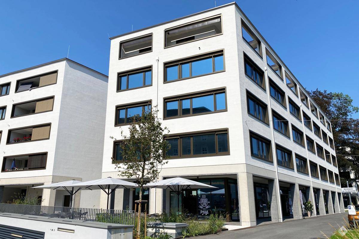 Modernes weißes Gebäude mit vielen Fenstern und klarem Himmel, umgeben von Grünflächen und Sitzgelegenheiten.