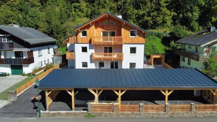 Luftaufnahme eines modernen Wohnhauses mit Holz-Carport und umgebender grüner Landschaft.
