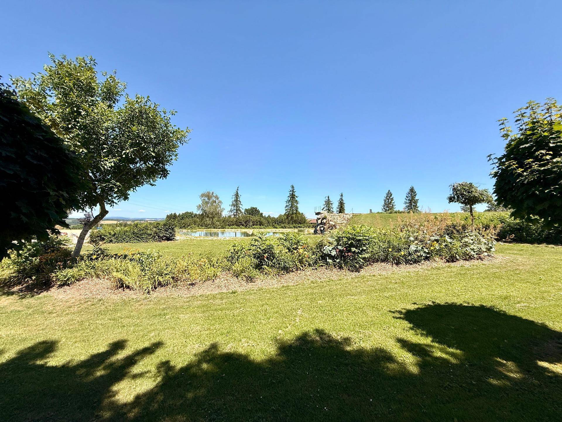 Üppiger grüner Gartenbereich mit Blumenbeeten und Blick auf einen Teich und die umliegende Landschaft.
