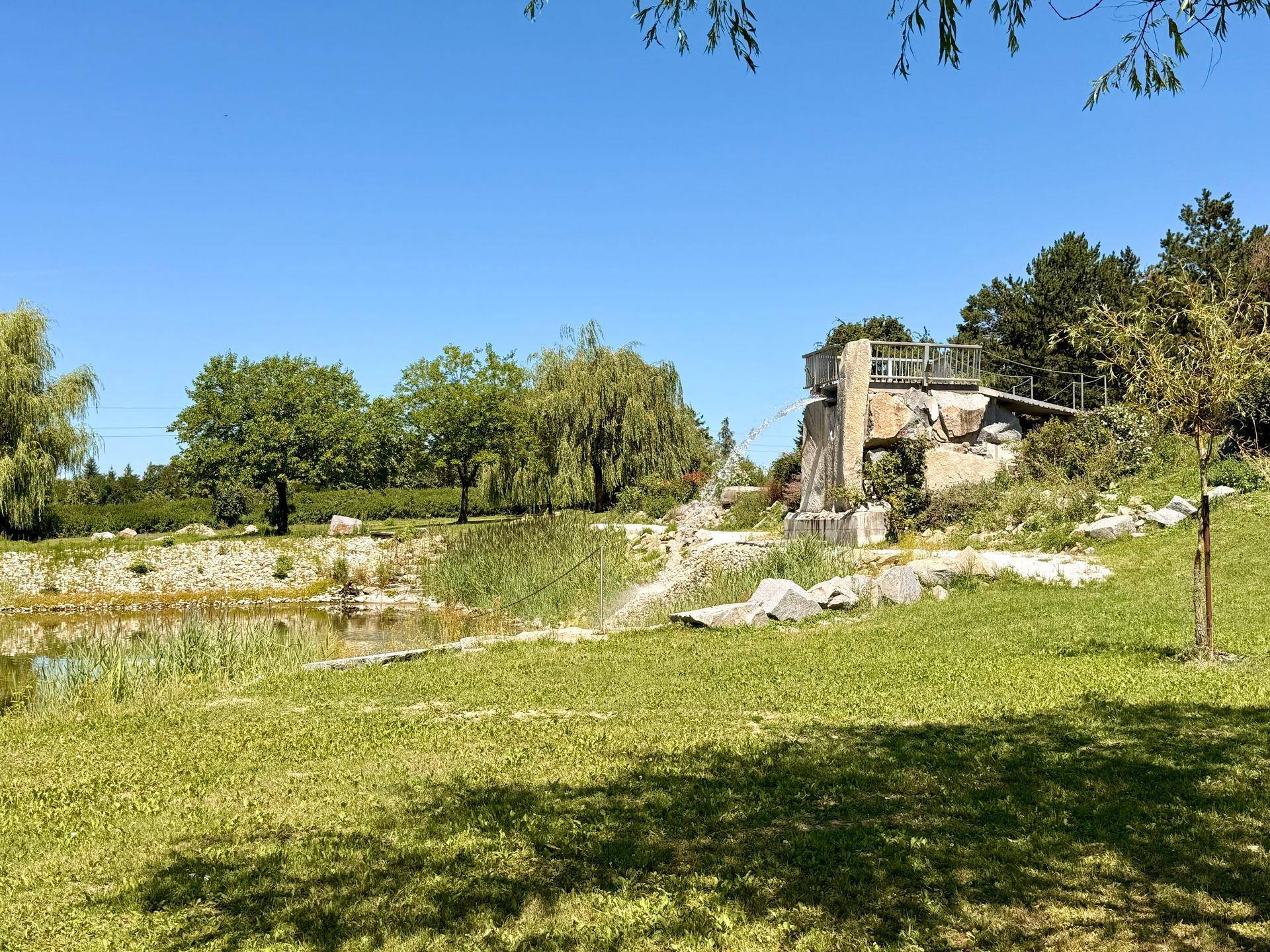 Natürlicher Teich mit dekorativem Wasserfall und kleiner Brücke, umgeben von Grünflächen und Bäumen.