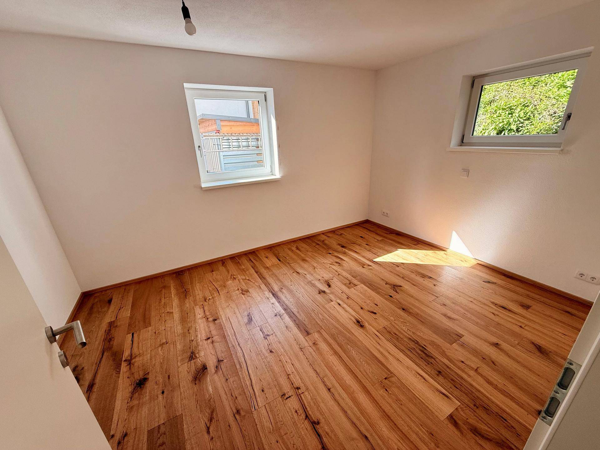 Leeres Schlafzimmer mit hellem Holzboden und zwei Fenstern, die viel Tageslicht hereinlassen.