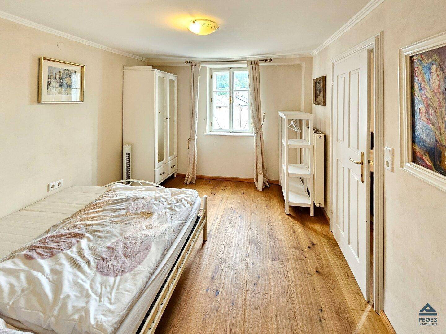 Helles Schlafzimmer mit Holzboden, weißem Kleiderschrank und einem Fenster mit Blick ins Grüne.