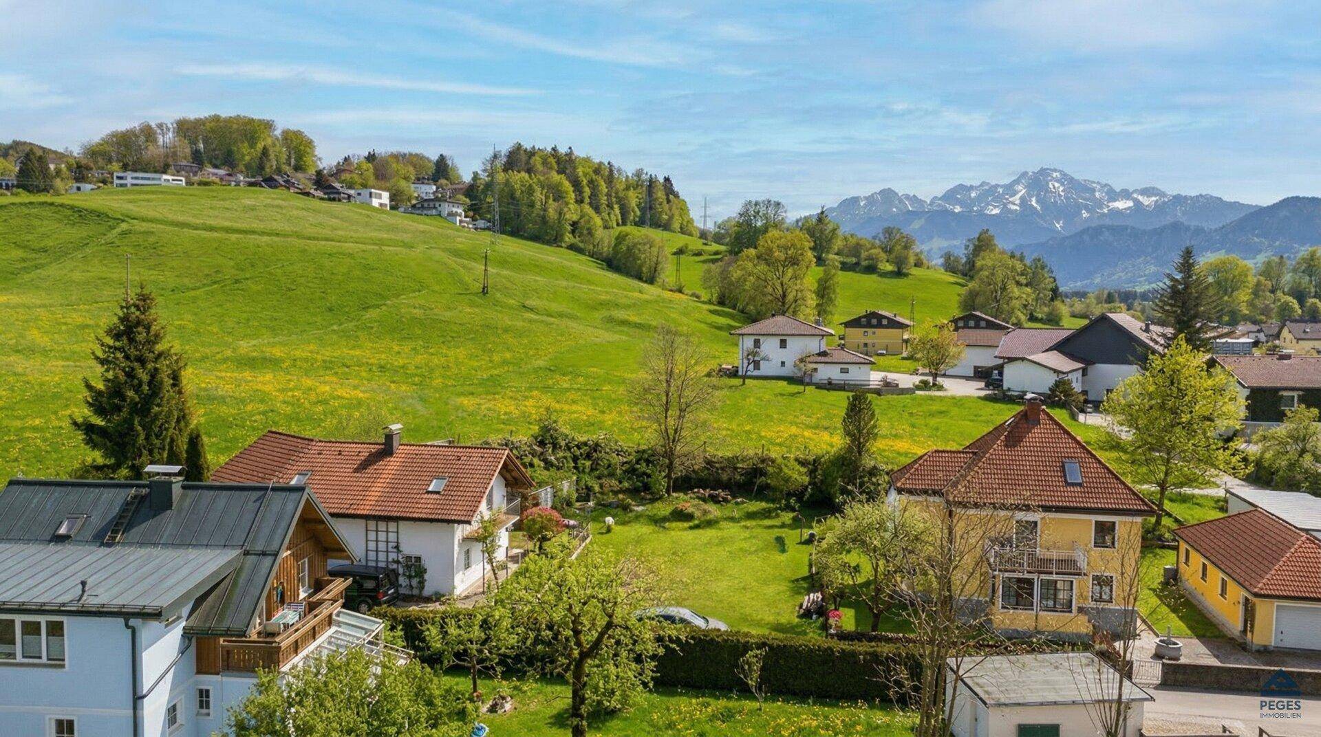 Weitläufige Landschaft mit Wohnhäusern auf sanften Hügeln und schneebedeckten Berggipfeln am Horizont.