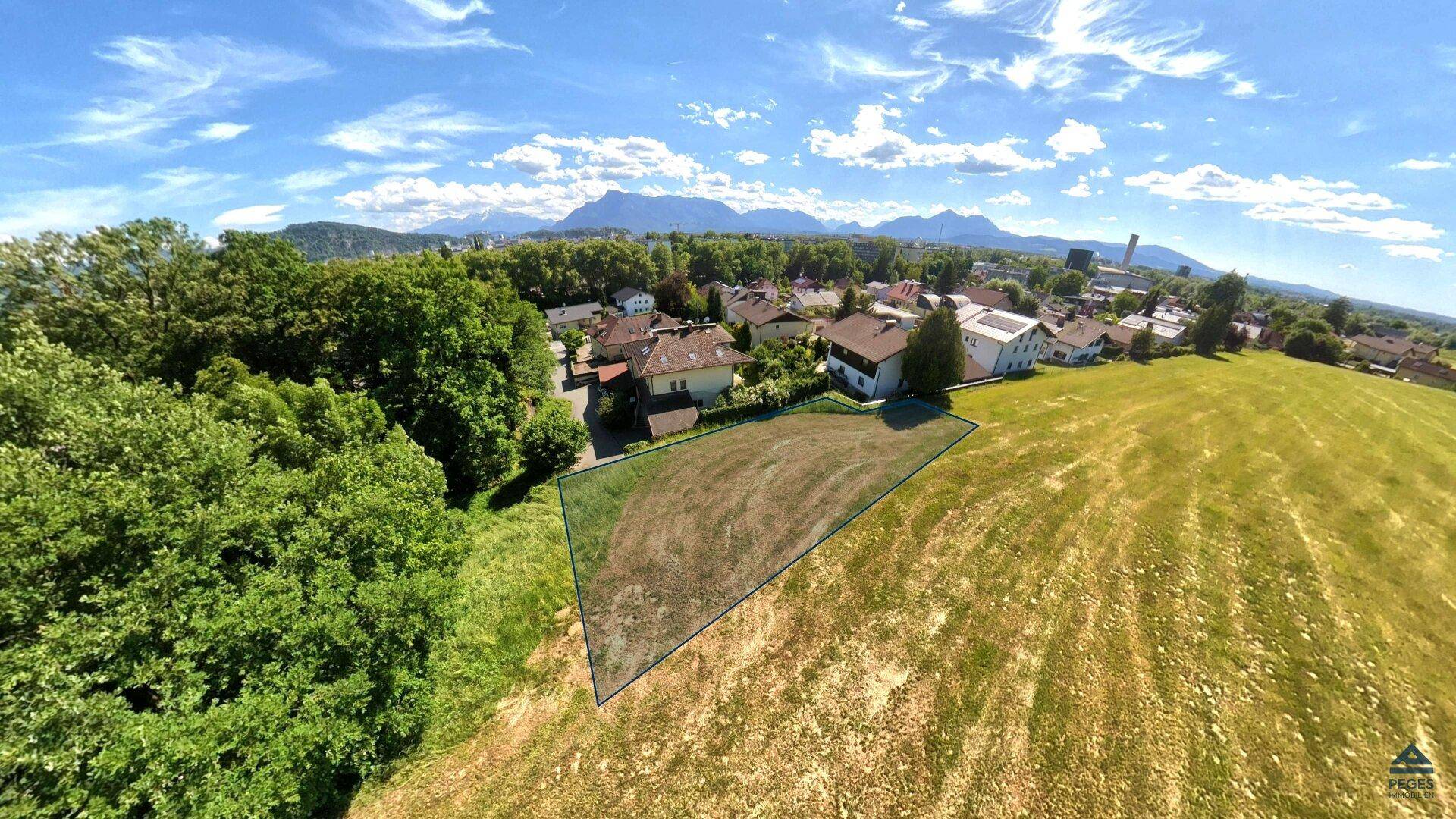 Luftaufnahme eines markierten Grundstücks mit Dorf und Alpenpanorama im Hintergrund unter blauem Himmel.