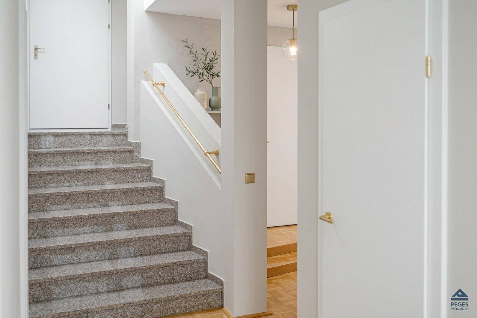 Heller Flur mit Steintreppe, goldenem Handlauf und weiß gestrichenen Wänden.