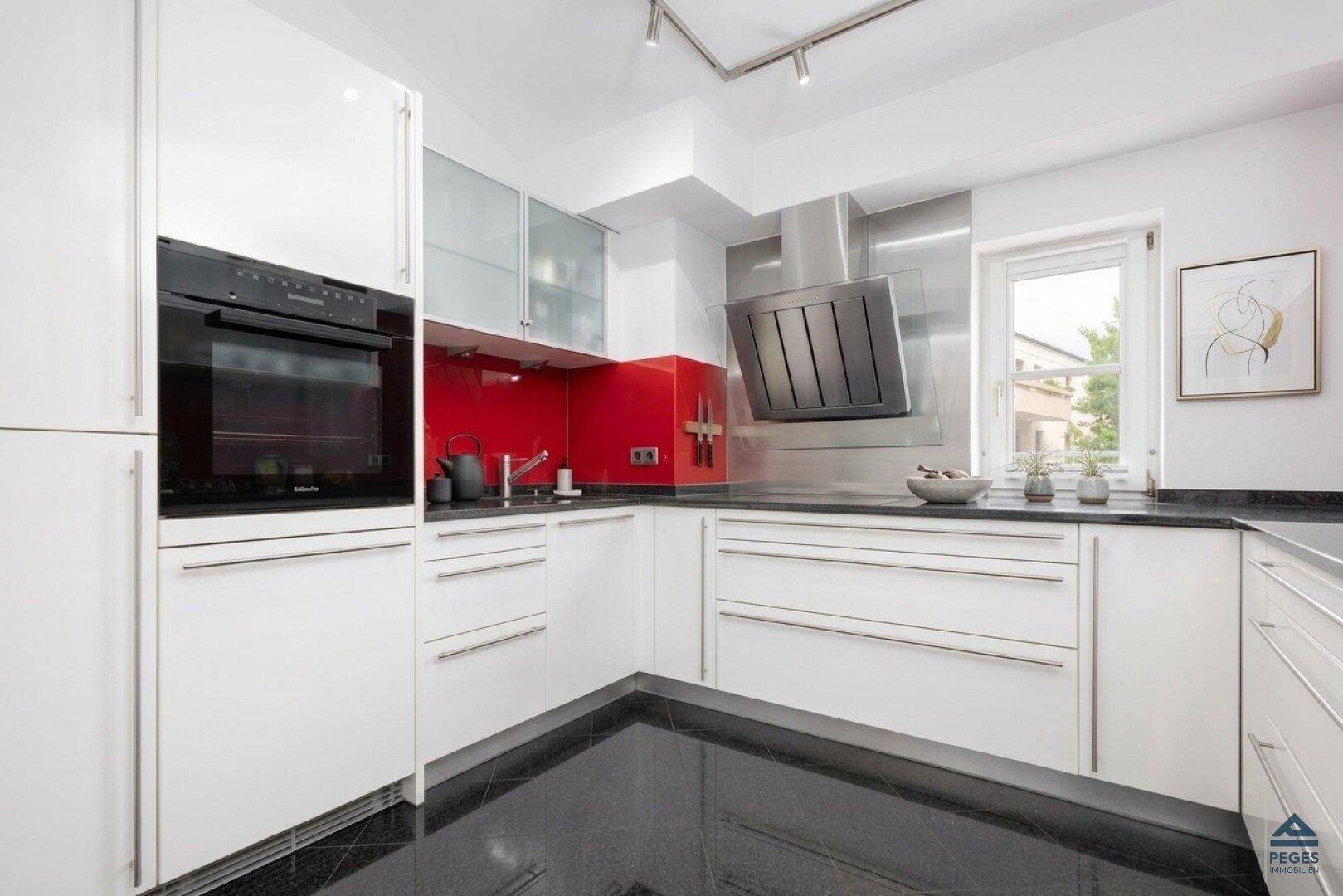 Moderne weiße Küche mit roter Rückwand, schwarzen Arbeitsplatten und integrierten Geräten.