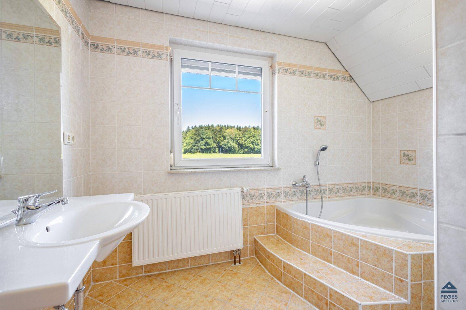 Badezimmer mit Badewanne, beigefarbenen Fliesen und Fenster mit Blick ins Grüne.