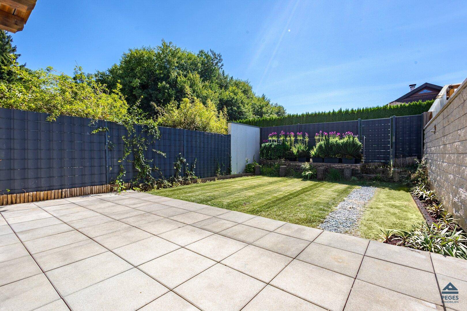 Gepflegter kleiner Garten mit Rasenfläche, gepflasterter Terrasse und grüner Hecke.
