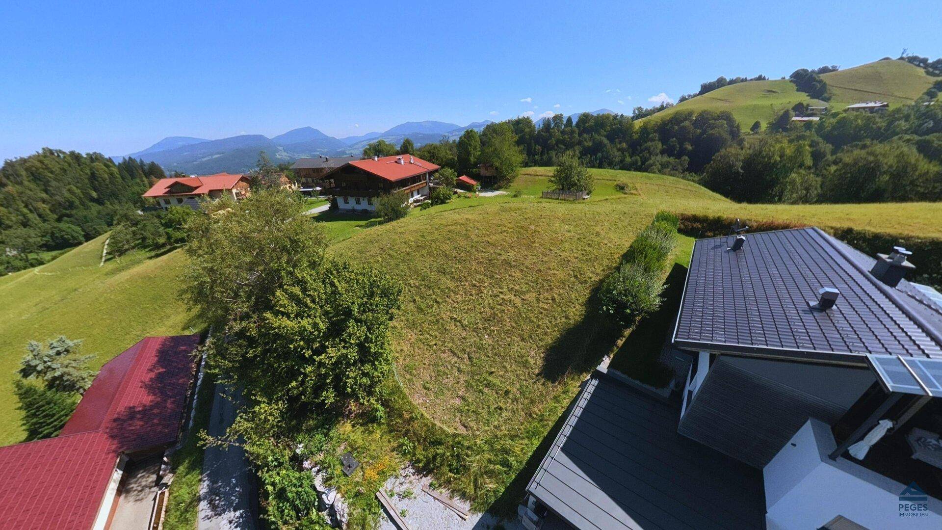 Blick über ein Hausdach hinweg auf eine weitläufige grüne Wiesenlandschaft mit bewaldeten Hügeln und Bergen.
