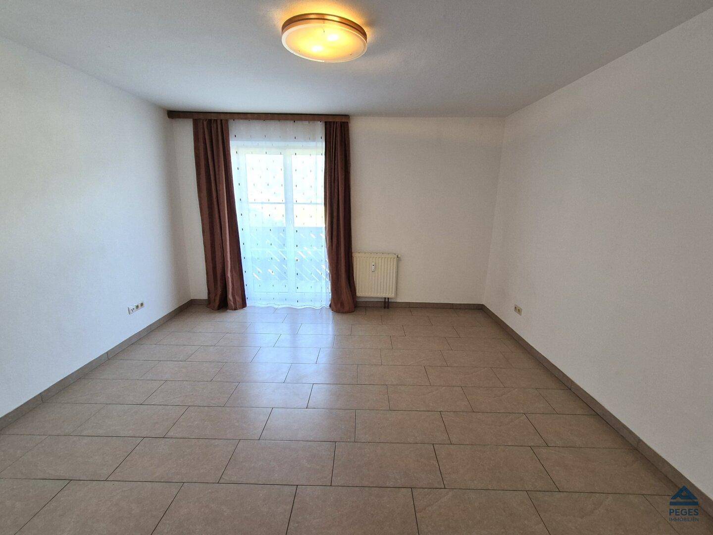 Leerer Raum mit hellen Fliesen, einem Fenster mit Vorhängen und einem Heizkörper.