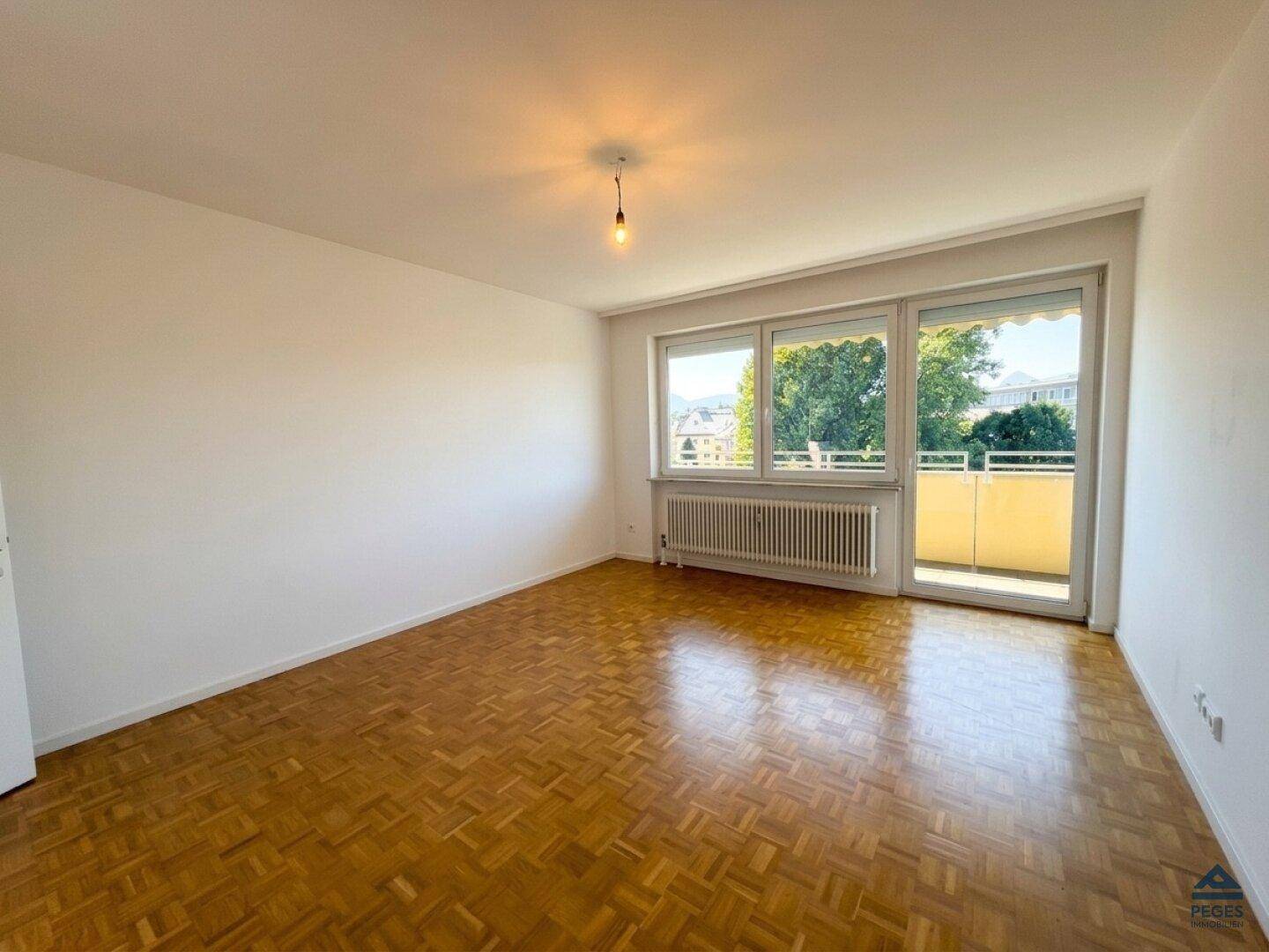 Helles Wohnzimmer mit Fischgrätparkettboden, großen Fenstern und Zugang zum Balkon.