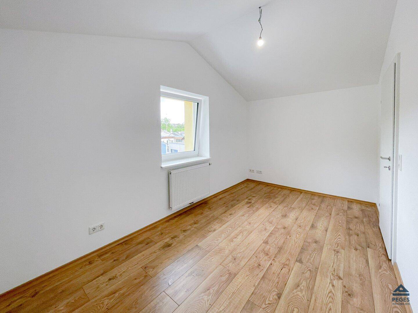 Helles Zimmer mit Holzboden, weiß gestrichenen Wänden und einem Fenster unter der Dachschräge.