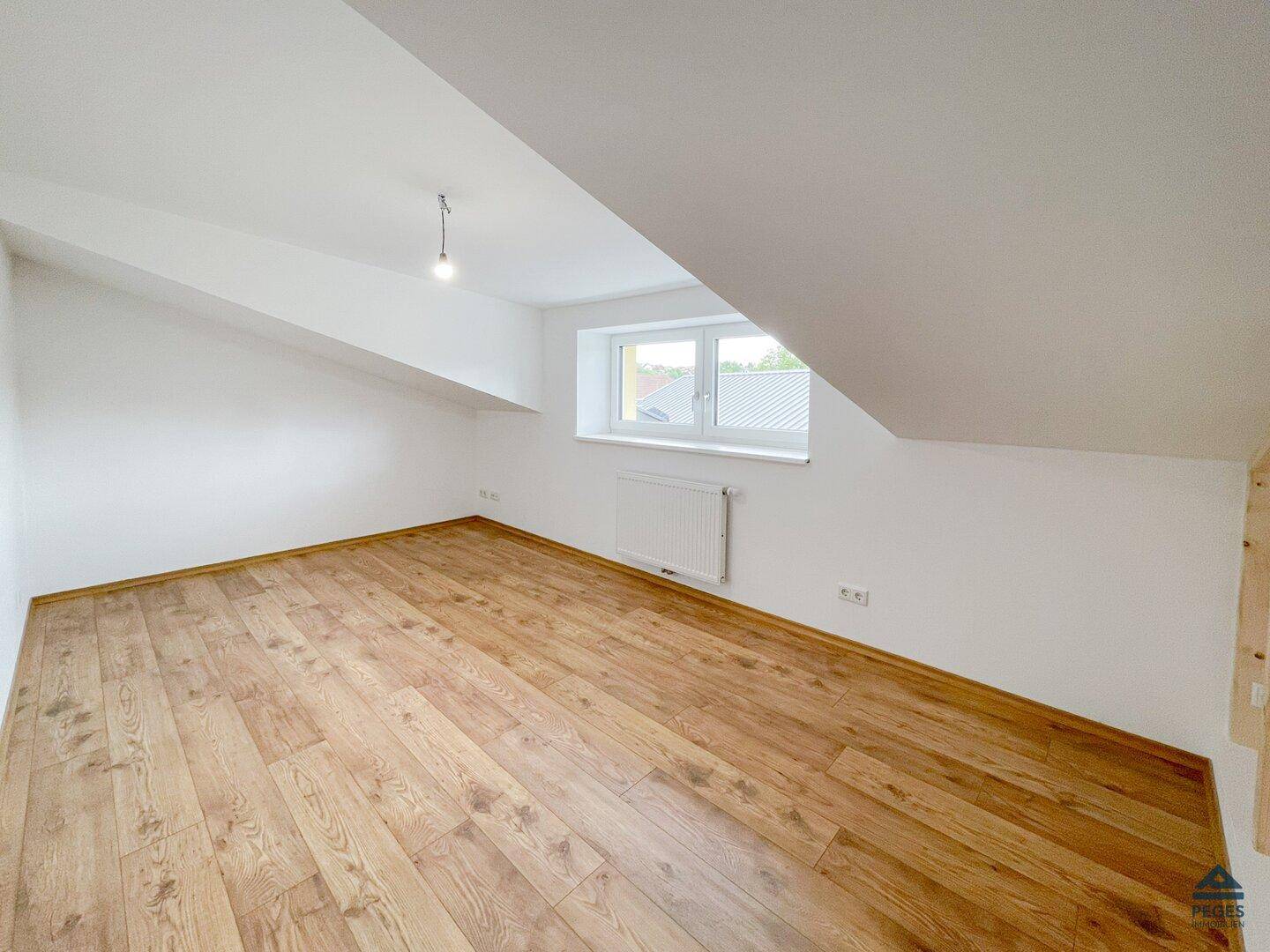 Weiteres helles Zimmer mit Holzboden, weißen Wänden und einem Fenster unter der Dachschräge.