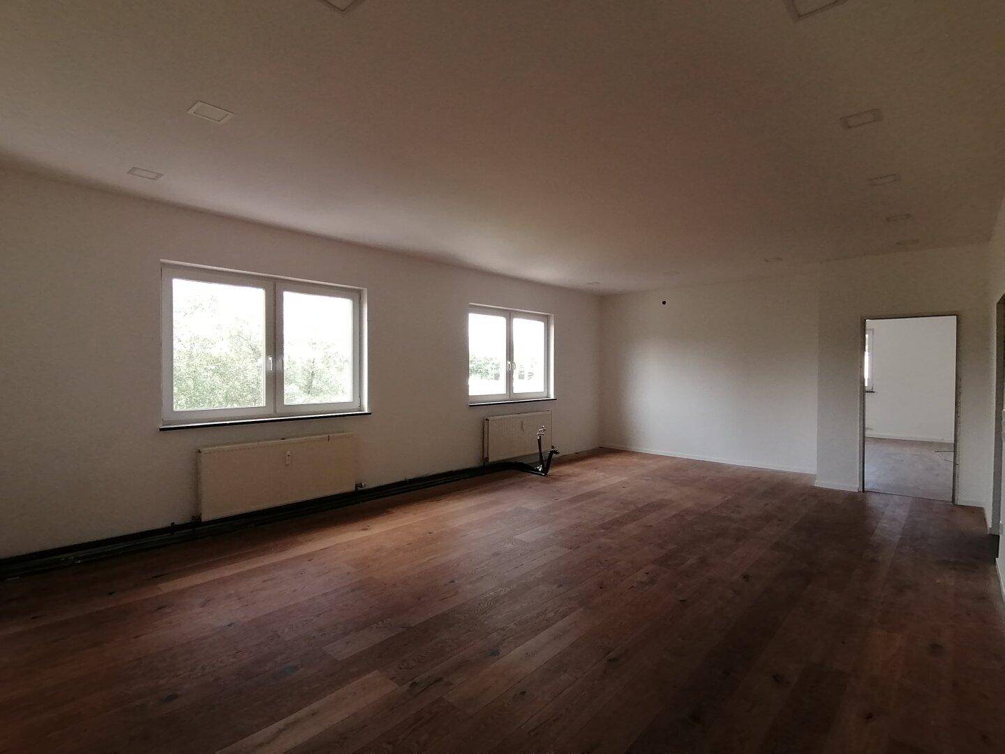 Geräumiger, leerer Raum mit Holzboden, zwei Fenstern und weiß gestrichenen Wänden.