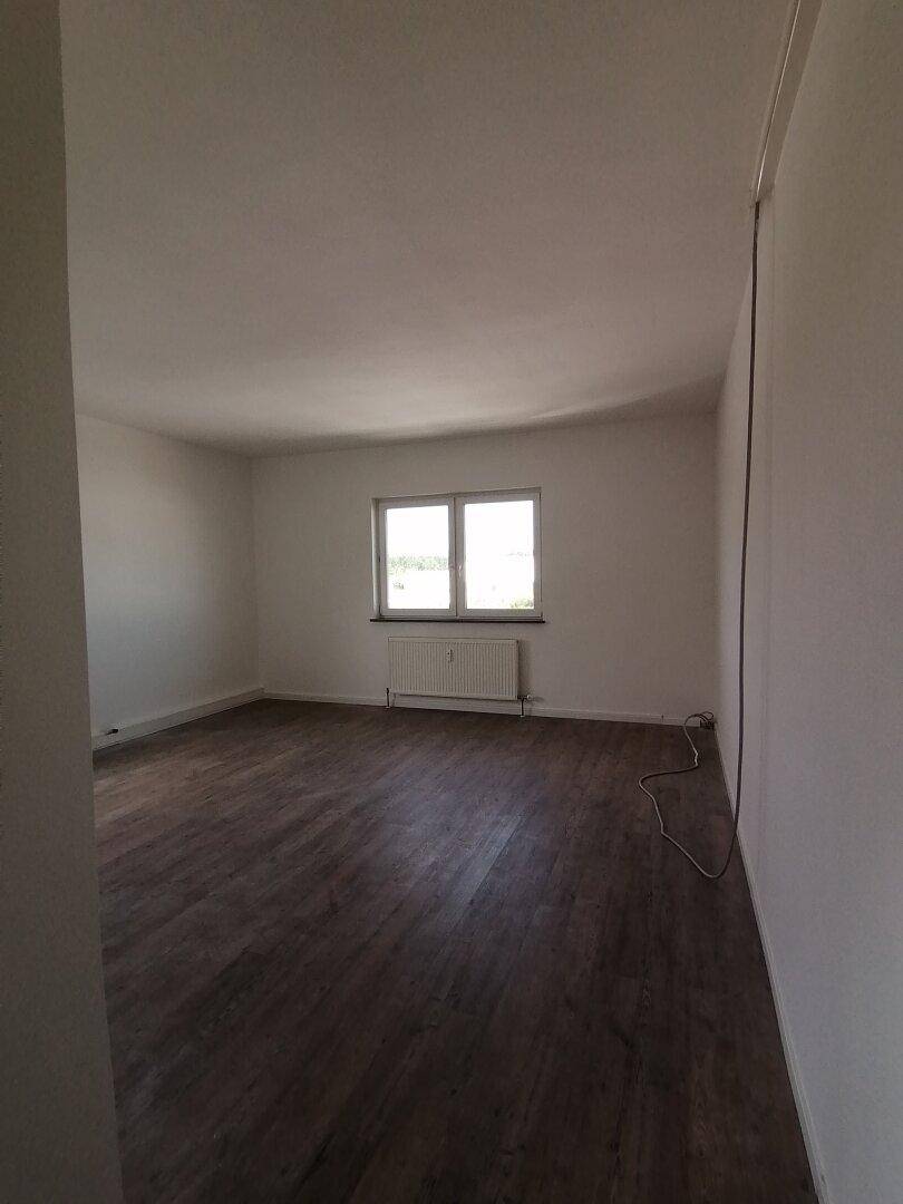 Leerer Raum mit dunklem Holzoptik-Boden und einem Fenster, das den Blick nach draußen freigibt.