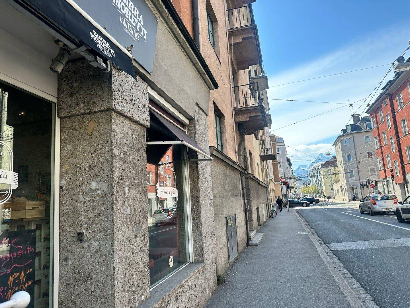 Seitenansicht eines Gebäudes mit Geschäften im Erdgeschoss und Balkonen in den oberen Etagen, entlang einer belebten Straße.
