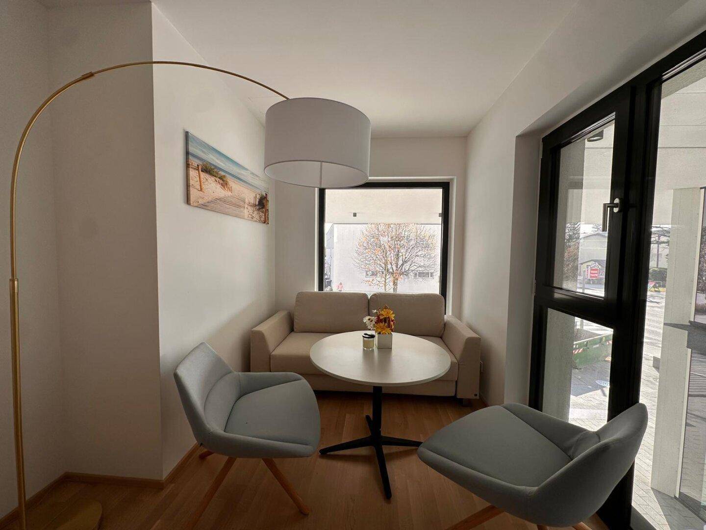Gemütlicher Wohnbereich mit beigem Sofa, zwei modernen Sesseln und viel Tageslicht durch große Fenster.