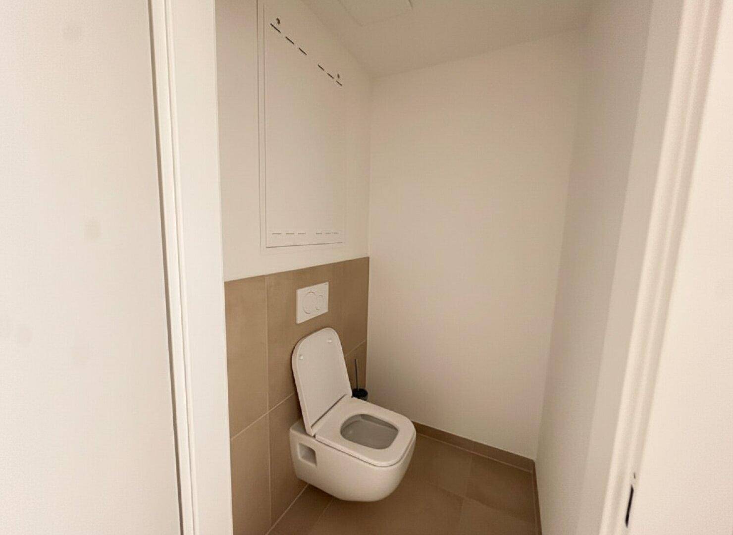 Kleine, saubere Gästetoilette mit wandmontiertem WC und beigefarbenen Fliesen im unteren Wandbereich.
