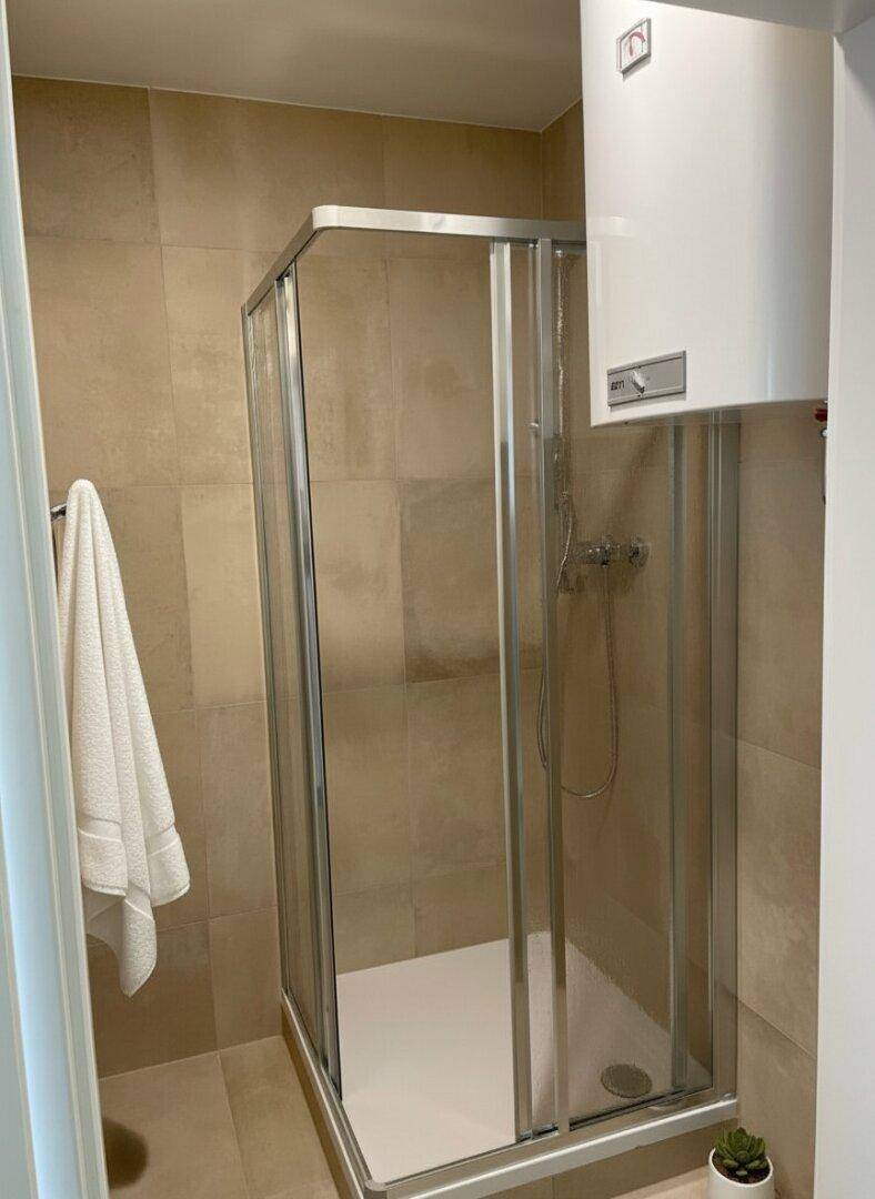 Modernes Badezimmer mit einer Duschkabine, beigefarbenen Fliesen und einem Warmwasserbereiter an der Wand.
