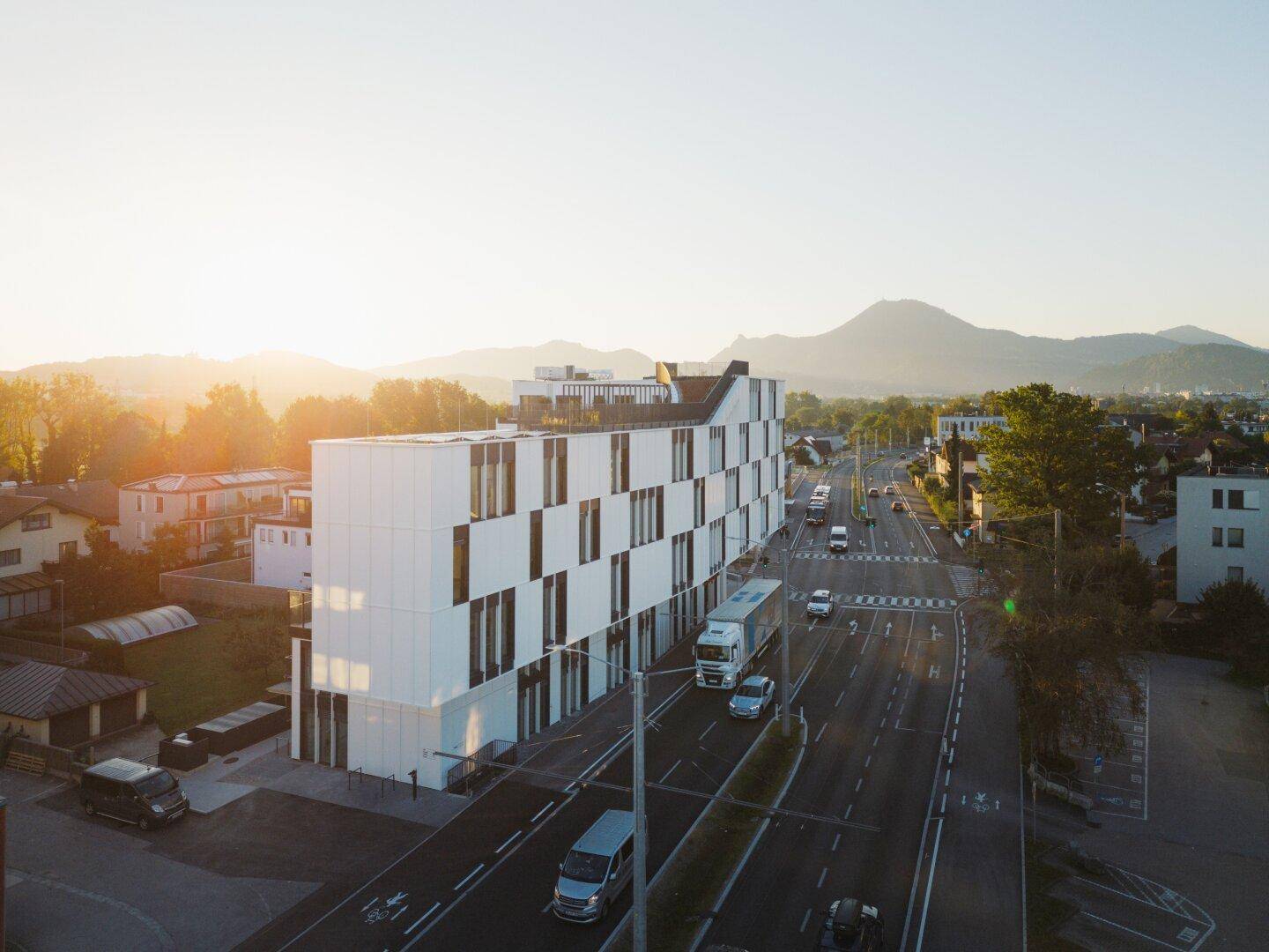 Moderne Architektur eines neuen Gebäudes an einer belebten Straße, umgeben von Bergen bei Sonnenuntergang.