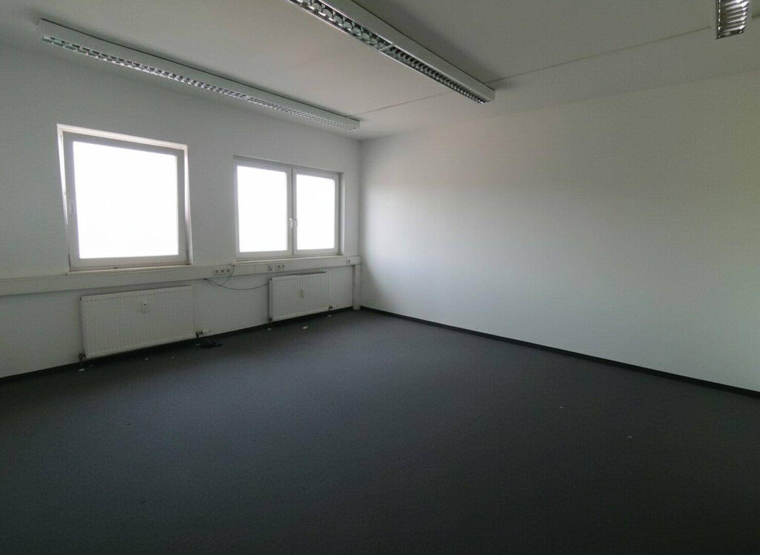 Geräumiges Büro mit Teppichboden, zwei Fenstern und funktionaler Deckenbeleuchtung.